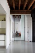 Blick durch raumhohe Glasschiebetür in restaurierter Loftwohnung mit rustikaler Holzbalkendecke und hellgrauem Kunstharzboden