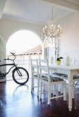 White dining table and refurbished second-hand chairs below chandelier; bicycle in background in front of open arched terrace doors