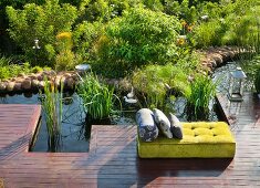 Gelbsamtenes Matrazenpolster mit grauen Kissen auf Holzdeck an idyllisch gestaltetem Wasserbecken mit Kiesel-Ufer und Schilfpflanzen