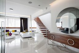 Eleganter, loftartiger Wohnraum - Designer-Sideboard unter rundem Spiegel an Wand, im Hintergrund Lounge in Weiß, auf hochglänzendem Boden
