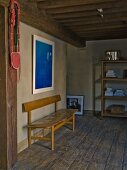 Simple, retro bench on rustic wooden floor in restored interior with wood-beamed ceiling and modern, blue artwork on wall