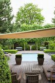 Sunshade over a wooden terrace, wooden outdoor furniture and plant containers in front of a pool