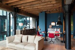 Sofa in open-plan interior of old factory floor with cast iron pillars and exposed roof beams; dining area in front of petrol blue back wall