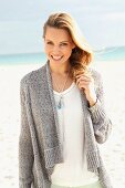 A blonde woman on a beach wearing a white blouse and a grey cardigan