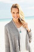 A blonde woman on a beach wearing a white blouse and a grey cardigan