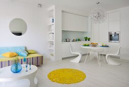 Lounge area, dining area and fitted kitchen in white, open-plan interior with round, yellow rug on white floor providing a splash of colour
