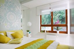 Bedroom with double bed, white and yellow bed linen and scatter cushions and wooden windows with white roller blinds and view of garden