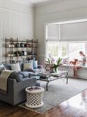 Sofa, coffee table, pale rug, woman seated on bay window seat and half-closed roller blinds