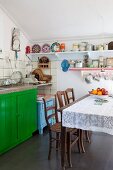 Dining table with lace tablecloth and antique chairs in front of sink on green-painted based unit in simple kitchen