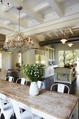 Dining area with classic chairs around rustic table, white jug of garden flowers and open-plan kitchen in background