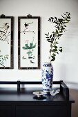 White and blue painted porcelain vase on black sideboard below framed pictures with Oriental motifs