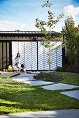 Sandspielplatz mit Kindern vor Garage mit Stahl-Plexiglas-Schiebetür in Zick-Zackmuster und sommerlichem Garten