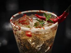 A champagne drink with chilli and ice cubes garnished with a chilli pepper and herbs in glass with a white chocolate rim