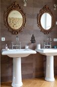 Twin pedestal sinks below mirrors with ornate, gilt frames on tiled wall