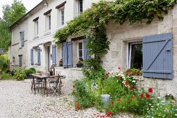 Mohnblumen vor rustikalem Landhaus mit blaugrau lackierten Fensterläden, Sitzplatz auf Kiesboden