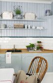 Pastel-blue, country-house kitchen with ornaments on floating shelves and Windsor chair at dining table in foreground