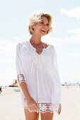 A blonde woman by the sea wearing a white blouse with a lace trim