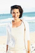 A dark-haired woman on a beach wearing a light t-shirt and jacket