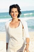 A dark-haired woman on a beach wearing a light t-shirt and jacket