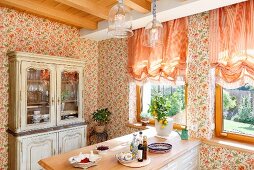Frühstückstheke in Küche mit Landhaus Charme, an Wand geblümte Tapete und apricotfarbene Raffrollos am Fenster