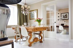 Various chairs around solid wooden dining table with carved base in traditional dining room with view into study through open double doors