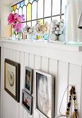 Pictures on white, wood-clad wall below stained-glass transom window with tealight holders and glass dishes on windowsill