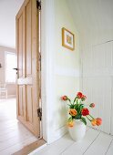 White vase of red and yellow tulips on white-painted wooden floor next to open interior door