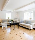 Pale sofas and white wicker chair around black coffee table in corner of rustic living room with whitewashed wood-beamed ceiling