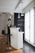 Chrome barstools at monolithic white counter below glass pendant lamps in front of French windows with white blinds and glasses on shelves