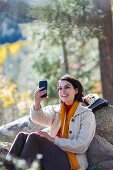 Frau in Wanderkleidung sitzt an Felsen gelehnt auf Waldlichtung & macht Selfie
