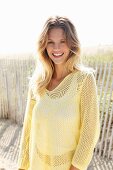 A young blonde woman on a beach wearing a white top and a pastel-yellow openwork jumper