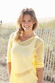 A young blonde woman on a beach wearing a white top and a pastel-yellow openwork jumper