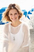 A young blonde woman on a beach wearing a white top and a transparent knitted jumper