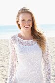 A young blonde woman on a beach wearing a white top and a transparent knitted jumper