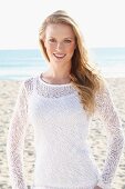 A young blonde woman on a beach wearing a white top and a transparent knitted jumper