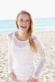 A young blonde woman on a beach wearing a white top and a transparent knitted jumper