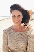 A brunette woman on a beach with her her blowing in the wind wearing a beige, openwork jumper