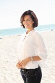 A brunette woman on a beach wearing a thin white blouse and jeans