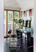 Modern metal chair in front of a desk with a glass top, in the corner of the room on a dark wooden floor, with French doors on the side with adjustable slat glass