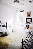 Yellow floral bedspread on black metal bed in front of small ladder-style shelves of ornaments