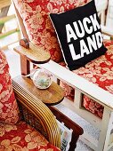 Black and white scatter cushion printed with 'Auckland' motto on floral armchair next to magazine rack
