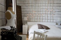 Simple bed with wooden headboard made of branches decorated with fairy lights, vintage typewriter and tailors' dummy in white, wood-clad bedroom