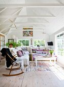 Rocking chair next to comfortable corner sofa with many scatter cushions in small summer house with white wood-clad interior