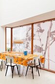 Retro chairs around table with orange tablecloth next to windows with roller blinds