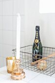 Goldfarbener Kerzenhalter mit weisser Kerze und Windlicht vor Maschendrahtkorb mit Champagnerflasche