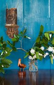 Geschnitzte Pelikanfigur und blühende Zweige in Vase, darüber Holz-Folklore an der lichtblau getönten Wand