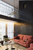 Traditionelles Sofa mit Brokatmuster an schwarzer Stuccolustro-Wand, davor Couchtisch aus filigranem, gebogenem Metall in modernem Wohnraum mit Galerie, Hängeleuchte im Fiftiestil