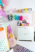 Toys on top of pale chest of drawers below string of colourful tassles and pictures on shelf