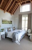 Double bed next to French windows with floor-length curtains in bedroom with exposed bamboo roof structure