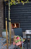 Herbstliche Terrasse mit dunklem Rattanstuhl, im Hintergrund Aussenkamin mit Feuer